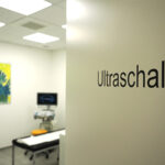 Halb geöffnete Tür mit Schriftzug Ultraschall, das Gerät und eine Liege im Hintergrund