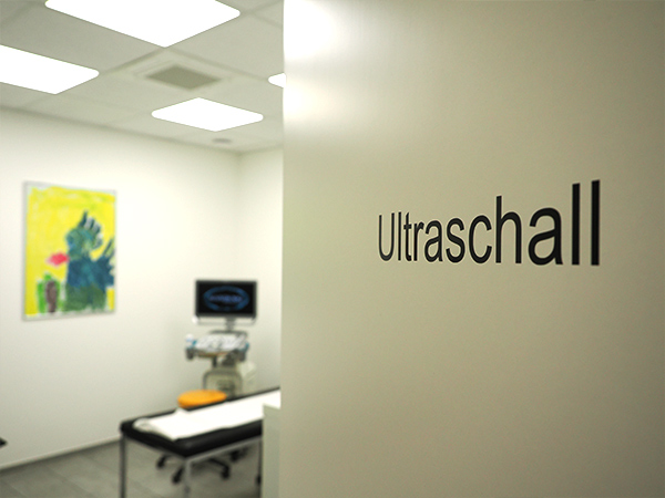 Halb geöffnete Tür mit Schriftzug Ultraschall, das Gerät und eine Liege im Hintergrund